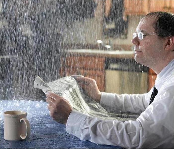 man reading newspaper with leak in ceiling. Man, table, and paper soaked.