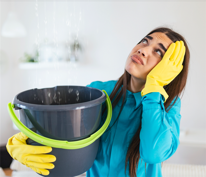 a woman in yellow gloves holding a bucket to catch water falling from her ceiling